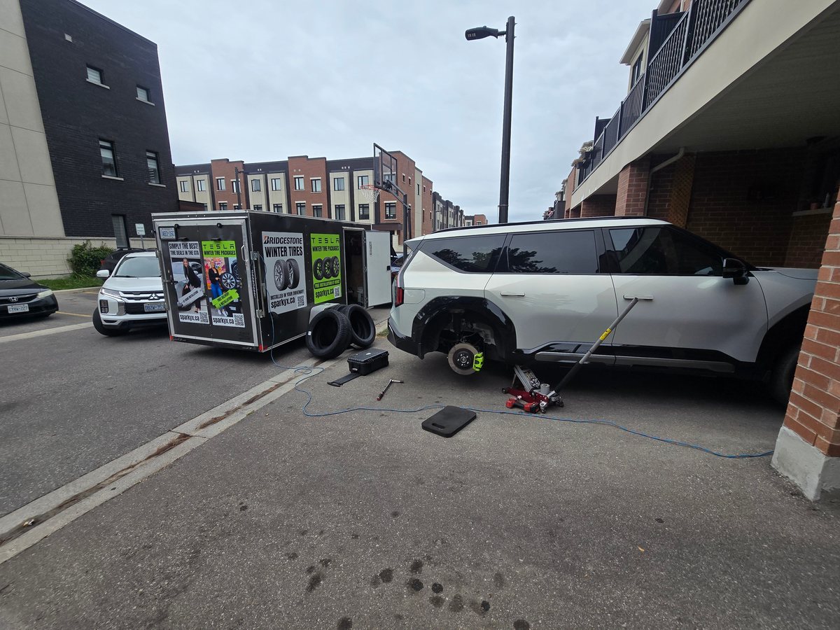 White Kia EV9 with its rear wheel removed during a driveway tire service, Sparky X trailer beside it